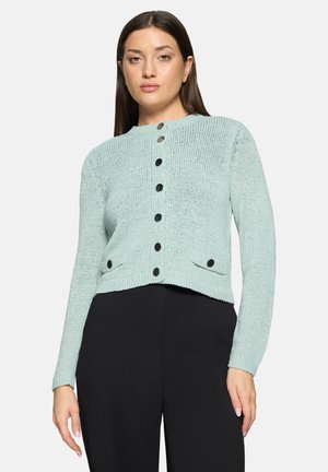 Femme portant un cardigan en maille bleu clair à boutons noirs et un pantalon noir, debout devant un fond uni.