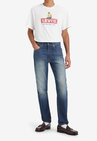 T-shirt blanc Levi's avec un logo rouge et un graphique de cowboy, associé à un jean droit bleu foncé. Le modèle porte des mocassins marron et des chaussettes blanches.