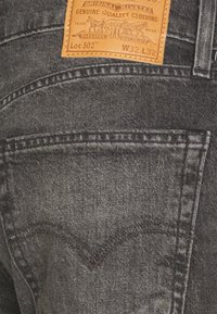 Jeans de mezclilla negra con un acabado texturizado, que presentan un parche de cuero trasero etiquetado como "Lote 502" en tono beige, y un diseño de costura curva clásico.