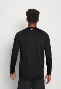 Czarna, długa koszulka sportowa z raglanowymi rękawami, o gładkiej teksturze i małym, białym logo na plecach przy karku. Prosty design.