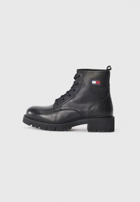 Bottines en cuir noir avec bout rond, lacets à l'avant, semelle en caoutchouc épaisse et un patch logo sur le côté. Texture lisse, design robuste.