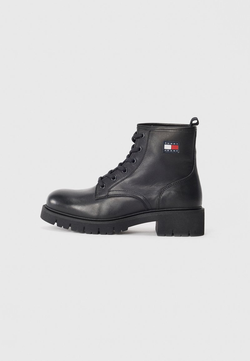 Bottines en cuir noir avec bout rond, lacets à l'avant, semelle en caoutchouc épaisse et un patch logo sur le côté. Texture lisse, design robuste.