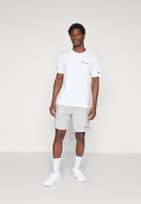 Hvid kortærmet T-shirt med et lille sort logo, parret med lysegrå shorts, hvide sneakers og hvide sokker, stående på en ensfarvet baggrund.