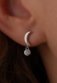 Silberne Ohrringe in Form eines halben Mondes mit einem hängenden runden Anhänger, der einen klaren Edelstein enthält. Das Design kombiniert geschwungene Formen mit polierter Textur.