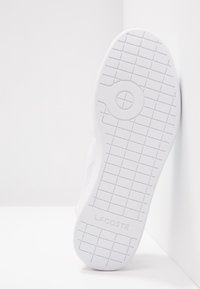 Semelle blanche de sneaker avec un motif en quadrillage et un motif circulaire près du centre, nom de la marque Lacoste embossé près du talon.