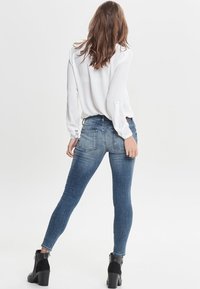 Jeans in denim skinny di un blu medio con leggere sfumature, abbinati a una blouse bianca a maniche lunghe e stivali neri alla caviglia.