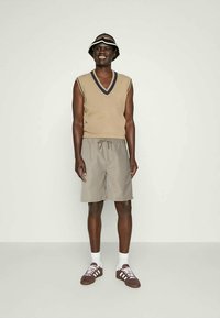 Smilende mann i en beige ermeløs genser, grå shorts med snøring, brune joggesko, hvite sokker og en svart-hvit bøttehatt.