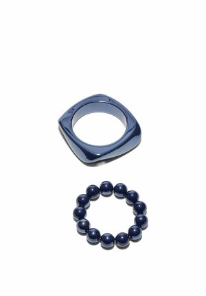 Bague carrée bleu foncé aux bords arrondis posée au-dessus d'un bracelet élastique assorti en perles bleu foncé sur fond blanc.