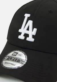 Schwarze Mütze aus Stoff mit einem weißen, gestickten "LA"-Logo. Enthält ein rundes silbernes Etikett mit der Aufschrift "New Era 9FIFTY Adjustable."