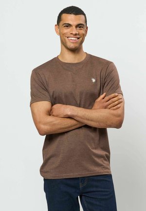 Lächelnder Mann mit kurzen Haaren, trägt ein braunes Kurzarmhemd und dunkelblaue Jeans, steht mit verschränkten Armen vor einem einfarbigen Hintergrund.