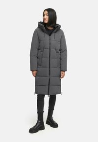 Grå puffjacka med huva, med dragkedja framtill och en strukturerad quiltad design. Bärs över svarta jeans och klumpiga svarta stövlar.
