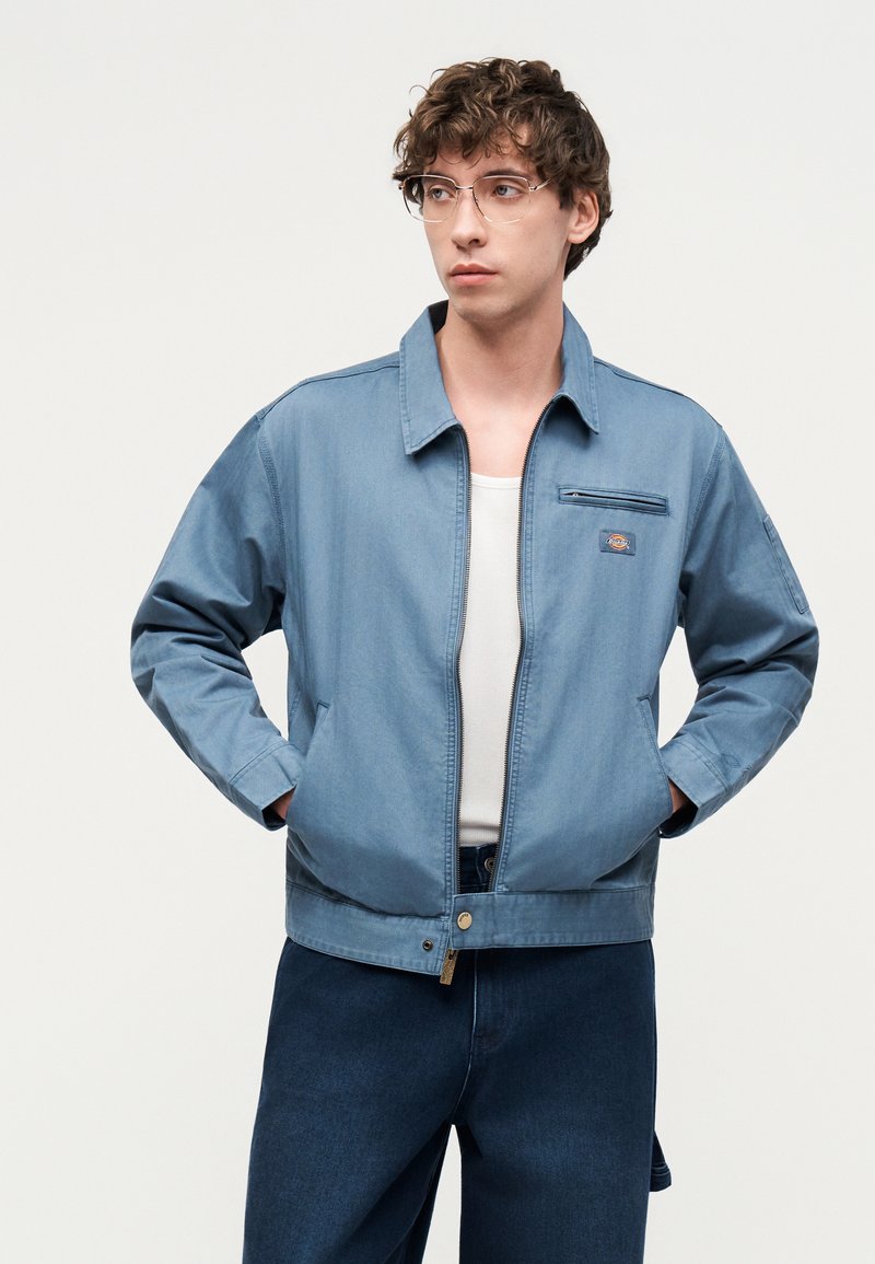 Jeune homme aux cheveux bouclés et portant des lunettes, vêtu d'une veste zippée bleu clair sur une chemise blanche et un pantalon bleu foncé, les mains dans les poches, regardant sur le côté.