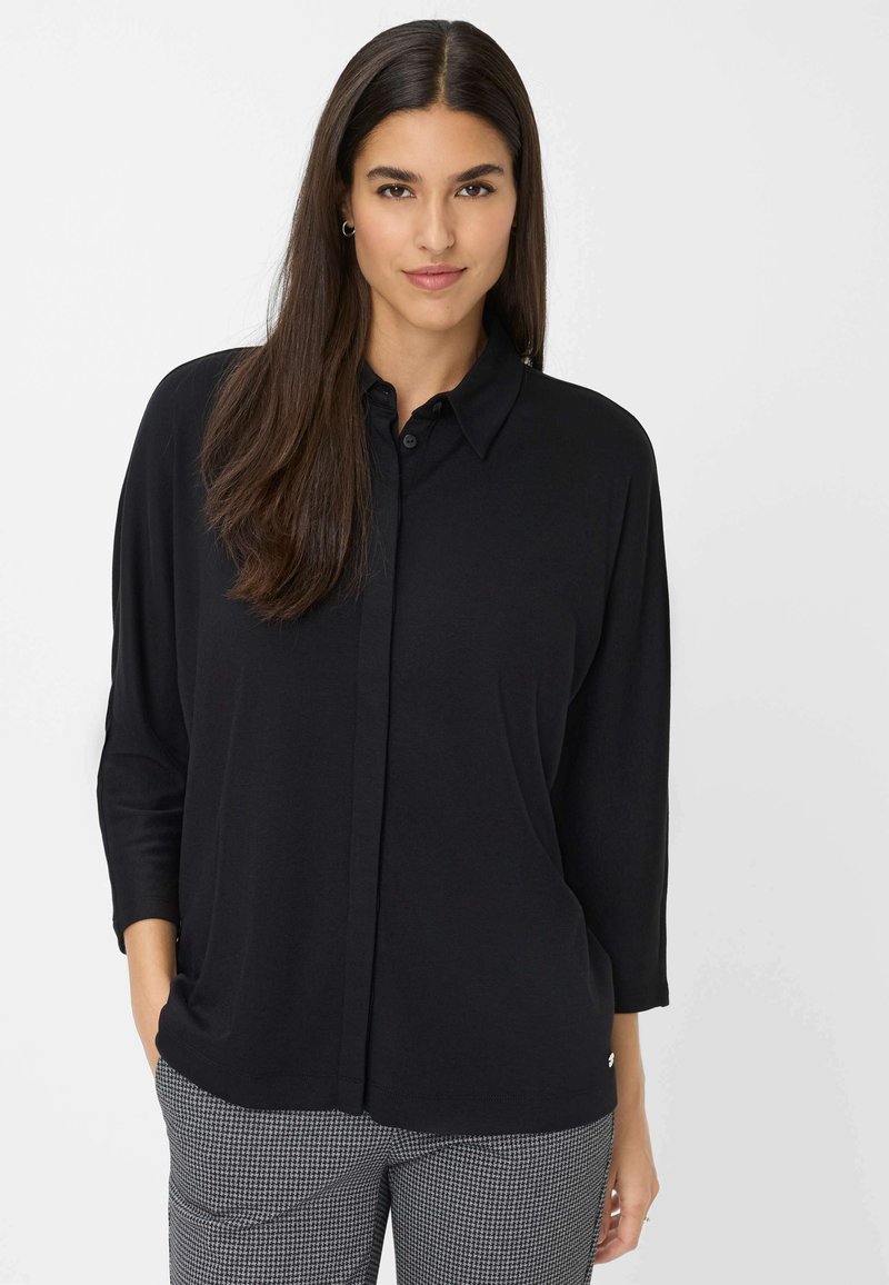 Zwarte blouse met lange mouwen, een kraag, knopen aan de voorkant en een ontspannen pasvorm. Gepaard met grijze broek met patroon in een houndstooth ontwerp.