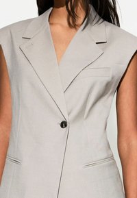 Ljusgrå ärmlös blazer med spetsiga slag, enkel knappstängning och sidofickor. Slät tygkvalitet med skräddarsydd passform och subtila detaljer.
