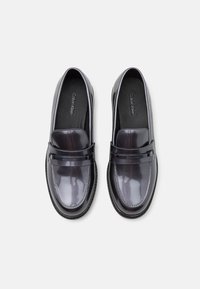 Mocassins en cuir verni noir avec design à enfiler, présentant un bout arrondi et une large sangle sur le dessus. Détails de semelle intérieure texturée visibles.