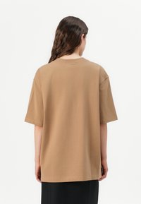 Smeđa oversized majica s kružnim izrezom, kratkim rukavima i glatkom pamučnom teksturom. Ima bočne proreze i opušteni kroj.