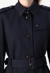 Marintrenchcoat med bälte i midjan, knäppning fram, axelklapp och skuren krage. Har mörkbruna knappar och en jämn textur.