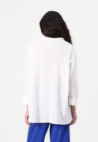 Valge linane pluus, millel on kaelus, pikad varrukad ja dekoratiivne helmed seljal. Kombineeritud siniste pükstega.