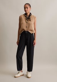 Beige knitted sleeveless top with ribbed accents, button detail, high-waisted black pants, and white sneakers with beige soles. Patterned scarf at neck.