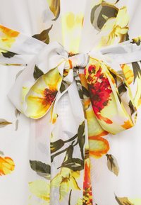 Tissu à motif bouquet avec des fleurs vibrantes jaunes, orange et rouges sur un fond clair ; il comporte un détail de taille nouée.