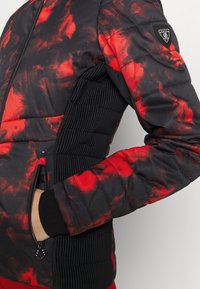 Doudoune noire et rouge à motif avec panneaux latéraux côtelés noirs, poche zippée et écusson sur la partie supérieure de la manche.
