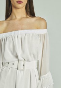 Weiße schulterfreie Bluse mit langen Ärmeln, gerafftem Stoff am Ausschnitt, versehen mit einem breiten Gürtel mit rechteckiger Schnalle.