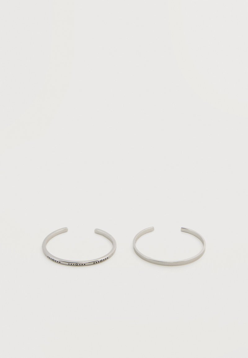 Δύο λεπτές ασημένιες βραχιόλια για μανσέτες, το ένα απλό και το άλλο χαραγμένο με Μπράιγ, σε ουδέτερο φόντο.