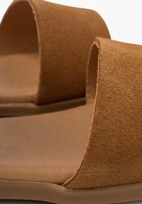 Hnědé semišové pantofle na skluz s širokými pásky a plochou gumovou podrážkou. Texturovaný povrch a čisté linie vytvářejí minimalistický design.