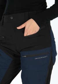 Zwarte en marineblauwe outdoorbroek met meerdere ritszakken, elastische tailleband en versterkte stiksels. Kenmerkend is het logo "SWEDEMOUNT."