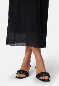 Des sandales noires à enfiler avec des tresses, dotées d'un bout carré et d'une finition mate texturée. Portées avec une jupe noire plissée.