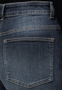 Gros plan sur la poche arrière d'un jean en denim bleu foncé avec des coutures visibles et des passants de ceinture sur fond noir.