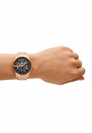 Roségoldene Uhr mit schwarzem Zifferblatt, das filigrane Zahnräder und silberne Stundenmarkierungen zeigt, auf einem passenden Armband im Stil eines Armbands.