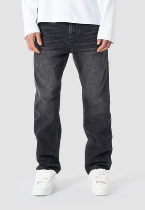 Person iført sorte vasket straight-leg jeans og hvide sneakers, stående med hænderne afslappet ned langs siden mod en ensfarvet baggrund.