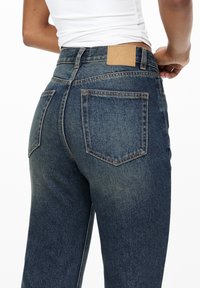 Dark blue denim jeans with a high waist, five pockets, and a tan leather label on the back waistband, featuring a slightly faded texture.