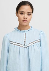 Blusa azzurro chiaro con un collo arricciato, maniche lunghe e dettagli in pizzo lungo il petto. Tessuto testurizzato con una vestibilità ampia.