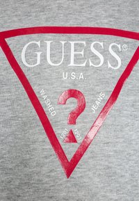 T-shirt in cotone grigio con grande design a triangolo rosso. Il testo include "GUESS U.S.A." e un punto interrogativo con dettagli "WASHED" e "JEANS".