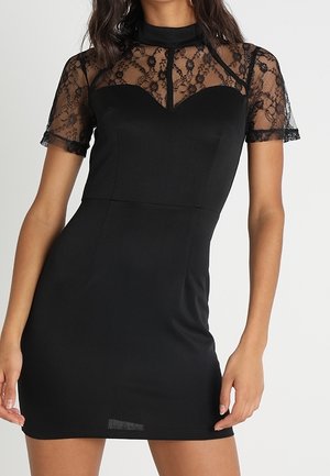 Femme portant une robe noire cintrée avec des manches courtes en dentelle transparente et un empiècement, un col montant et un détail corsage cœur.