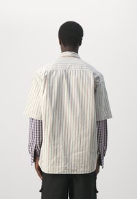 Mężczyzna odwrócony plecami, ubrany w luźną, paskowaną koszulę z krótkim rękawem na fioletowej, w kratkę koszuli z długim rękawem oraz czarne spodnie, na jednolitym tle.