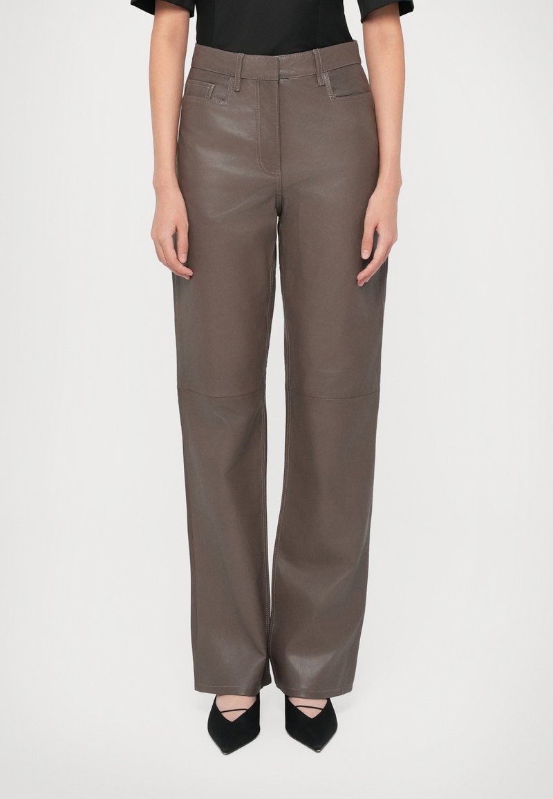 Vrouw in bruin leren broek met hoge taille en rechte pijpen, voorzien van voorzakken en zwarte puntige schoenen met enkelschoenen.