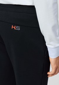 Zwarte sweatpants met een gladde textuur, voorzien van een oranje en grijze "HS" logo op de linker heup, gecombineerd met een lichtblauwe mouw.