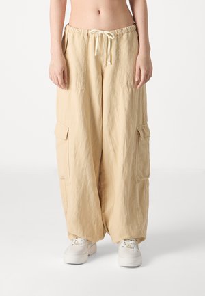 Persoon draagt beige cargo-broek met wijde pijpen en grote zakken, gecombineerd met witte sneakers, staand tegen een effen lichte achtergrond.