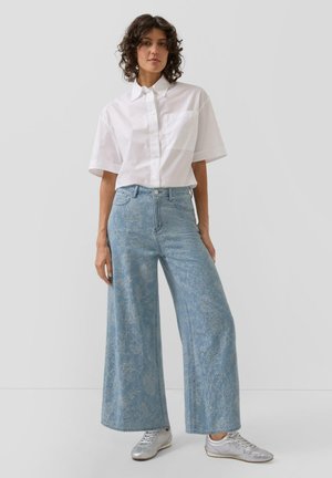 Vrouw staand, gekleed in een wit overhemd met korte mouwen, hooggetaillede brede blauwe jeans met bloemenpatroon en zilveren sneakers.