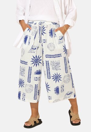 Løse hvide bukser med blå mønstre af sol, muslingeskaller og blade, kombineret med beige sandaler og hænder i lommerne, båret sammen med en hvid skjorte og top.