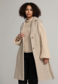 Beige wollen blend jas met een uitgesneden kraag, twee knopen en zijzakken; gecombineerd met een gestructureerde beige gebreide trui en zwarte broek.