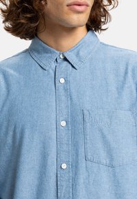 Camisa de botones azul claro hecha de tela de mezclilla, con un collar puntiagudo, botones blancos y un bolsillo en el pecho. Superficie suave y texturizada.