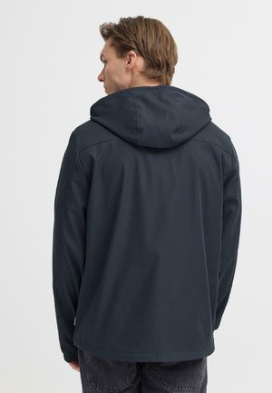 Mann, von hinten, trägt eine schwarze Kapuzenjacke mit elastischen Bündchen und dunkle Jeans vor einem einfarbigen hellen Hintergrund.
