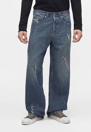 Person trägt lockere, ausgewaschene blaue Jeans mit ausgefransten Stellen und graue Sneakers, steht vor einem schlichten hellen Hintergrund.