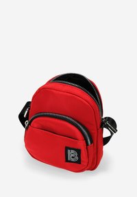 Mochila roja de nylon con dos compartimentos con cremallera, acentos negros y un destacado parche de logo con la letra "B". Diseño compacto y redondeado con una correa para el hombro.