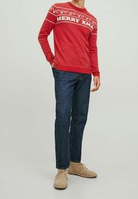 Roter gestrickter Pullover mit dem Text "MERRY XMA", weißen Mustern und Punkten. Kombiniert mit dunkelblauen Jeans und hellbeigen Schuhen.