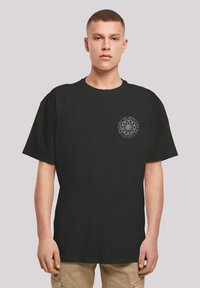 Schwarzes Baumwoll-T-Shirt mit kurzen Ärmeln. Verfügt über ein kreisförmiges grafisches Design in Grau auf der linken Brust. Lockere Passform, Rundhalsausschnitt.